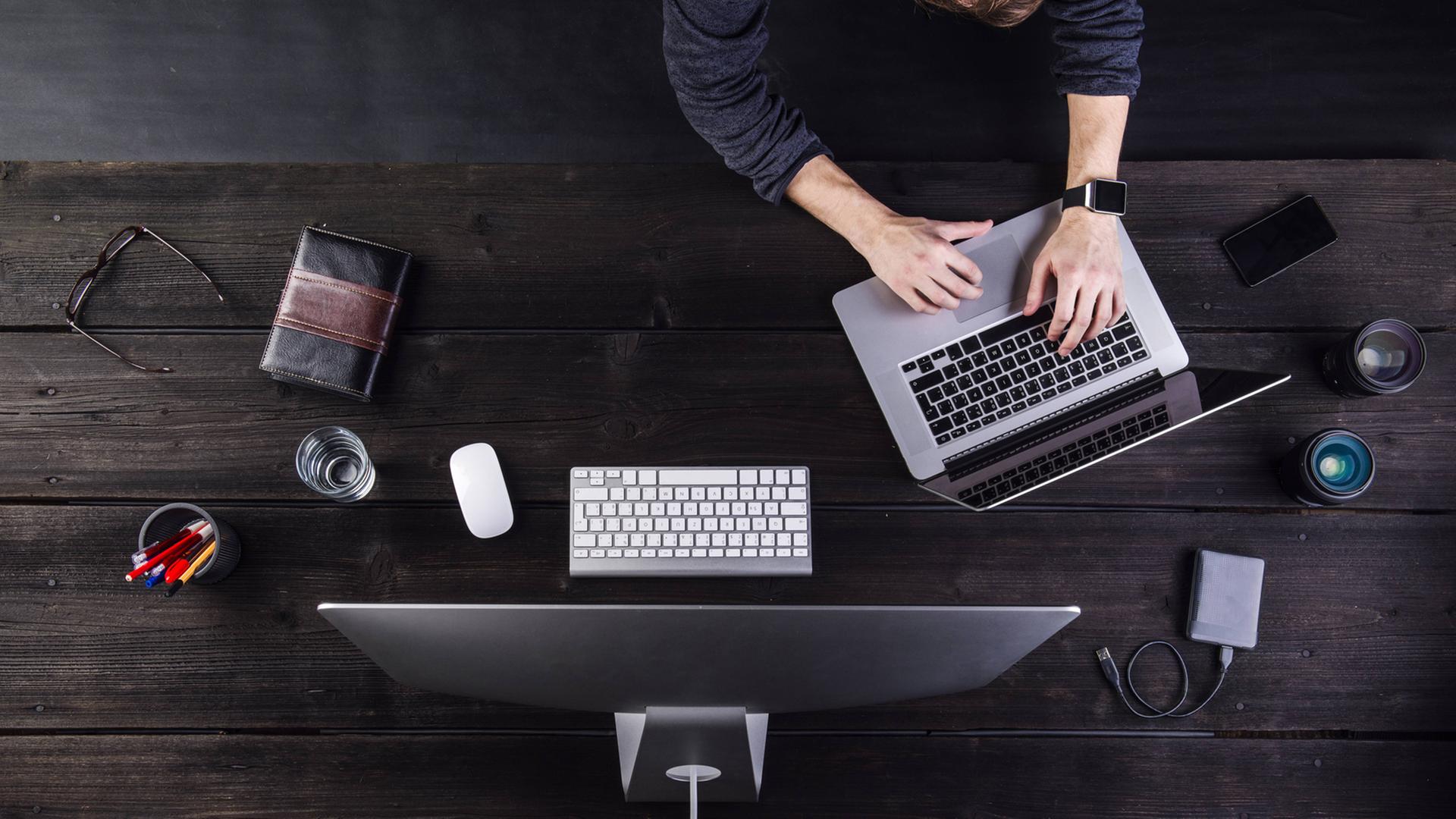 Ein Mann ist von oben zu sehen, wie er an einem Tisch mit dem Computer und mehreren anderen digitalen Geräten wie Notebook, Smartphone, Smartwatch arbeitet. Ein Mann ist von oben zu sehen, wie er an einem Tisch mit dem Computer und mehreren anderen digitalen Geräten wie Notebook, Smartphone, Smartwatch arbeitet.
