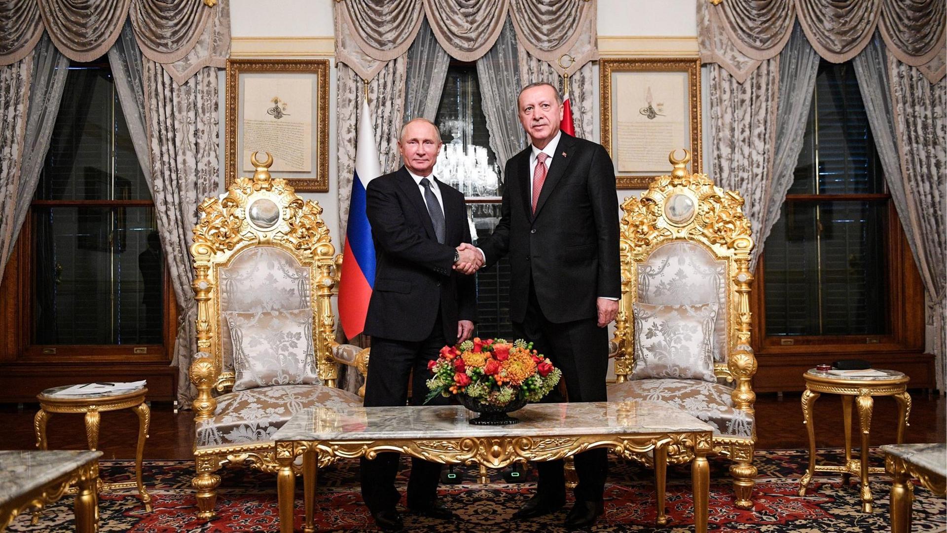 Putin und Erdogan stehen in einem pompös eingerichteten Raum hinter einem Tisch und zwischen zwei mit Gold verzierten Stühlen und schütteln sich die Hände. Putin und Erdogan stehen in einem pompös eingerichteten Raum hinter einem Tisch und zwischen zwei mit Gold verzierten Stühlen und schütteln sich die Hände.