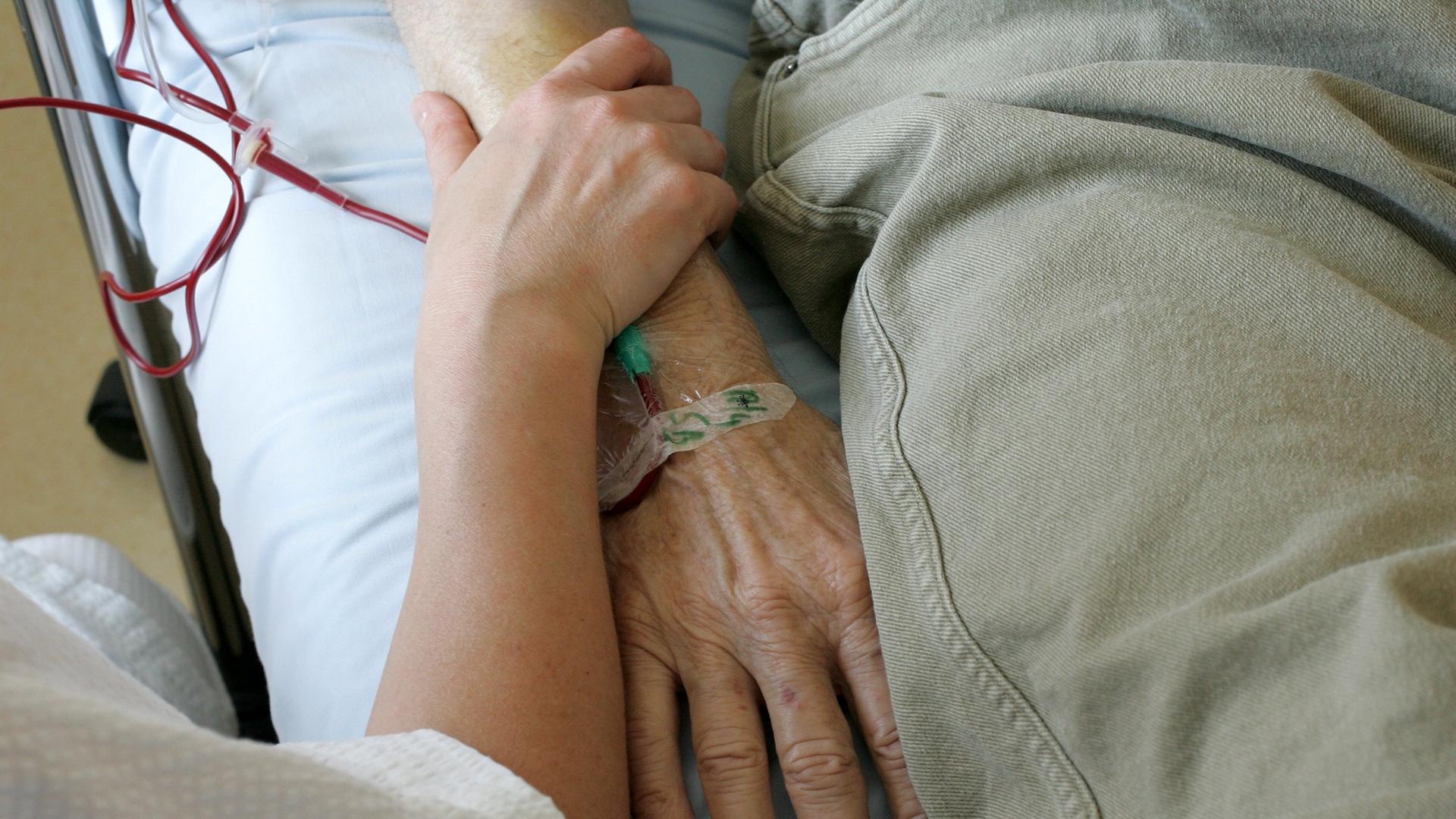 Ein jüngerer Mensch umfasst das Armgelenk einer älteren Person, die im Krankenbett liegt. Ein jüngerer Mensch umfasst das Armgelenk einer älteren Person, die im Krankenbett liegt.
