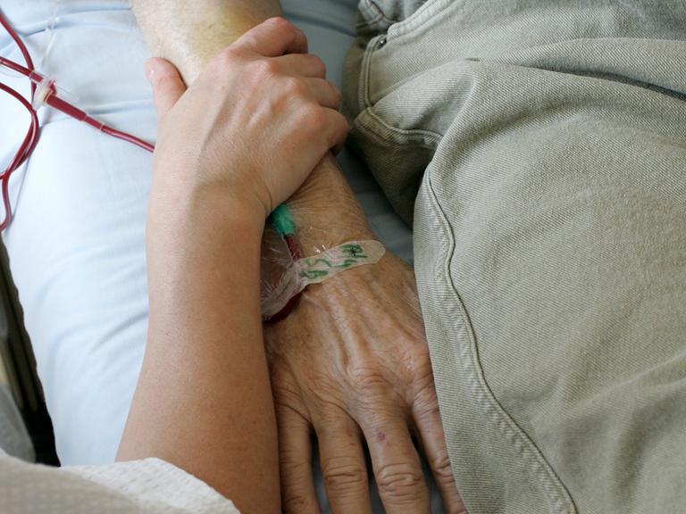 Ein jüngerer Mensch umfasst das Armgelenk einer älteren Person, die im Krankenbett liegt.