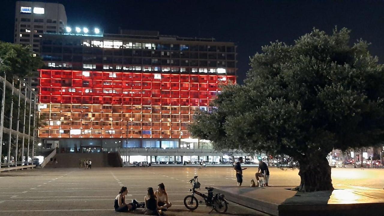 Blick in der Nacht auf das Rathaus, dessen Stockwerke von oben nach unten schwarz-rot-gold beleuchtet sind. Davor ein leerer Parkplatz mit einigen Menschen. Blick in der Nacht auf das Rathaus, dessen Stockwerke von oben nach unten schwarz-rot-gold beleuchtet sind. Davor ein leerer Parkplatz mit einigen Menschen.