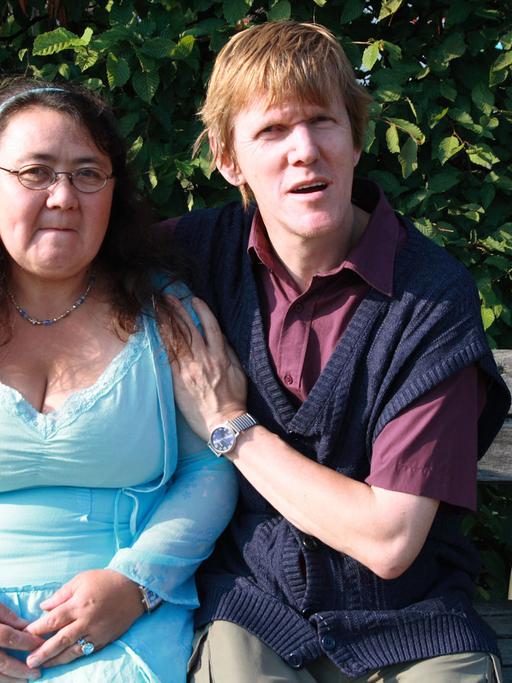 Elsa und Olaf lernten sich über eine Partnervermittlung für Menschen mit Behinderung kennen. Ein junges Paar sitzt auf einer Bank und schaut in die Kamera. Im Hintergrund Bäume von einem Park