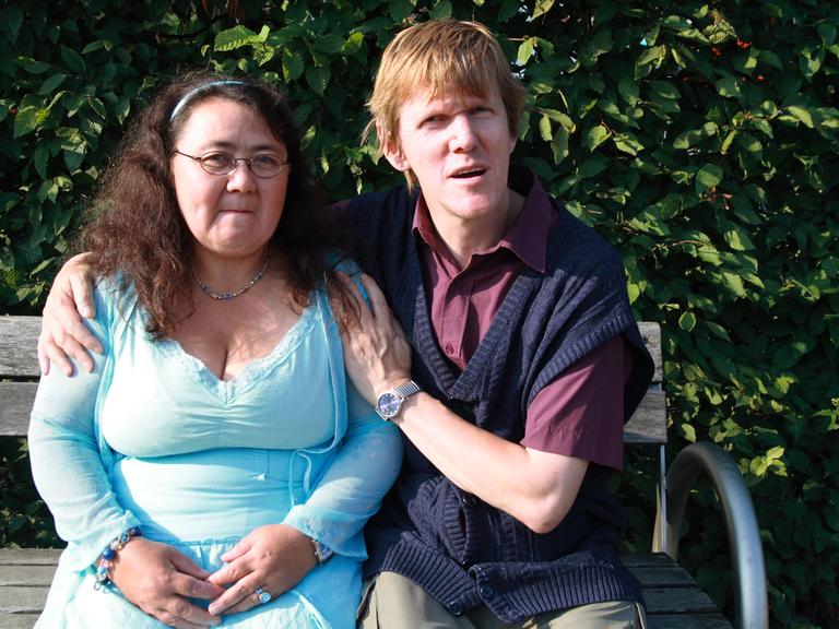 Elsa und Olaf lernten sich über eine Partnervermittlung für Menschen mit Behinderung kennen. Ein junges Paar sitzt auf einer Bank und schaut in die Kamera. Im Hintergrund Bäume von einem Park