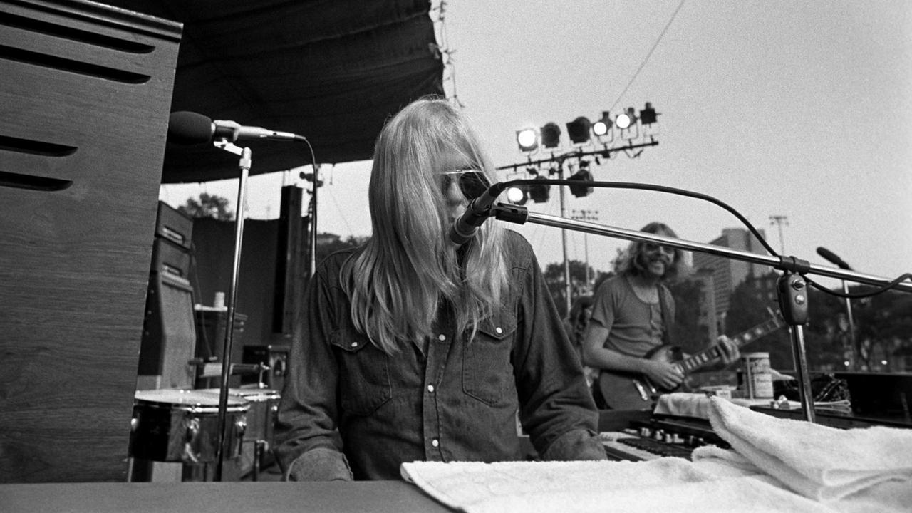 Ein Mann mit langen blonden Haaren sitzt an einer Orgel und singt in ein Mikrofon, im Hintergrund grinst sein Kollege an der Gitarre.