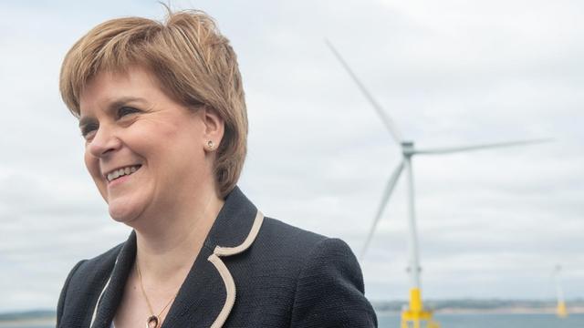 Schottlands Erste Ministerin Nicola Sturgeon vor blauem Himmel, im Hintergrund ist ein Windrad in der Nordsee zu sehen