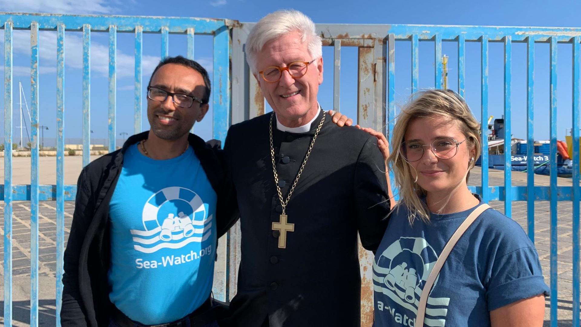Heinrich Bedford-Strohm, der Ratsvorsitzende der Evangelischen Kirche in Deutschland (EKD, M), steht mit Sea-Watch-Sprecherin Giorgia Linardi (rechts) und Sea-Watch-Kapitän Arturo Centore im Hafen. Das Schiff «Sea-Watch 3» liegt hier im JUni 2019 im Hafen. freigegeben. Foto: Annette Reuther/dpa | Heinrich Bedford-Strohm, der Ratsvorsitzende der Evangelischen Kirche in Deutschland (EKD, M), steht mit Sea-Watch-Sprecherin Giorgia Linardi (rechts) und Sea-Watch-Kapitän Arturo Centore im Hafen. Das Schiff «Sea-Watch 3» liegt hier im JUni 2019 im Hafen. freigegeben. Foto: Annette Reuther/dpa |