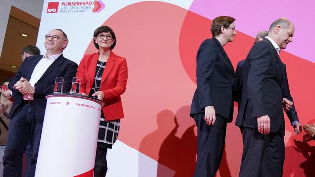 Norbert Walter-Borjans und Saskia Esken stehen vor dem SPD-Parteizeichen auf der Bühne, während das unterlegene Kandidaten-Duo (Olaf Scholz und Klara Geywitz) abtritt.