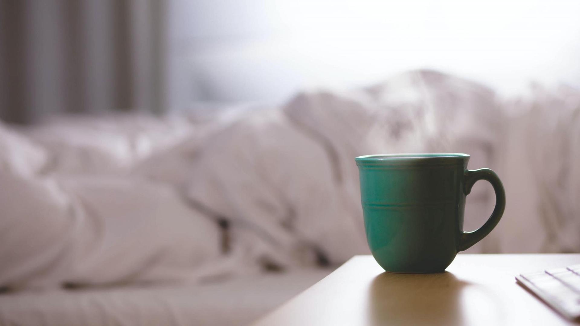 Eine Tasse mit heißem Getränk steht auf einem Tisch neben einem ungemachten Bett. Eine Tasse mit heißem Getränk steht auf einem Tisch neben einem ungemachten Bett.