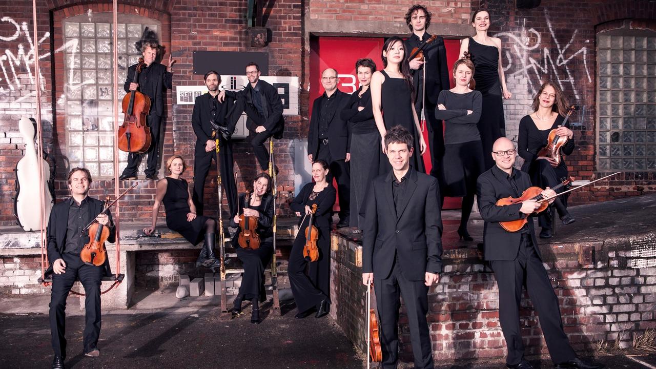 Schwarz gekleidete Musiker in Abendgarderobe stehen und sitzen mit ihren Instrumenten vor einem Fabrikgebäude Schwarz gekleidete Musiker in Abendgarderobe stehen und sitzen mit ihren Instrumenten vor einem Fabrikgebäude