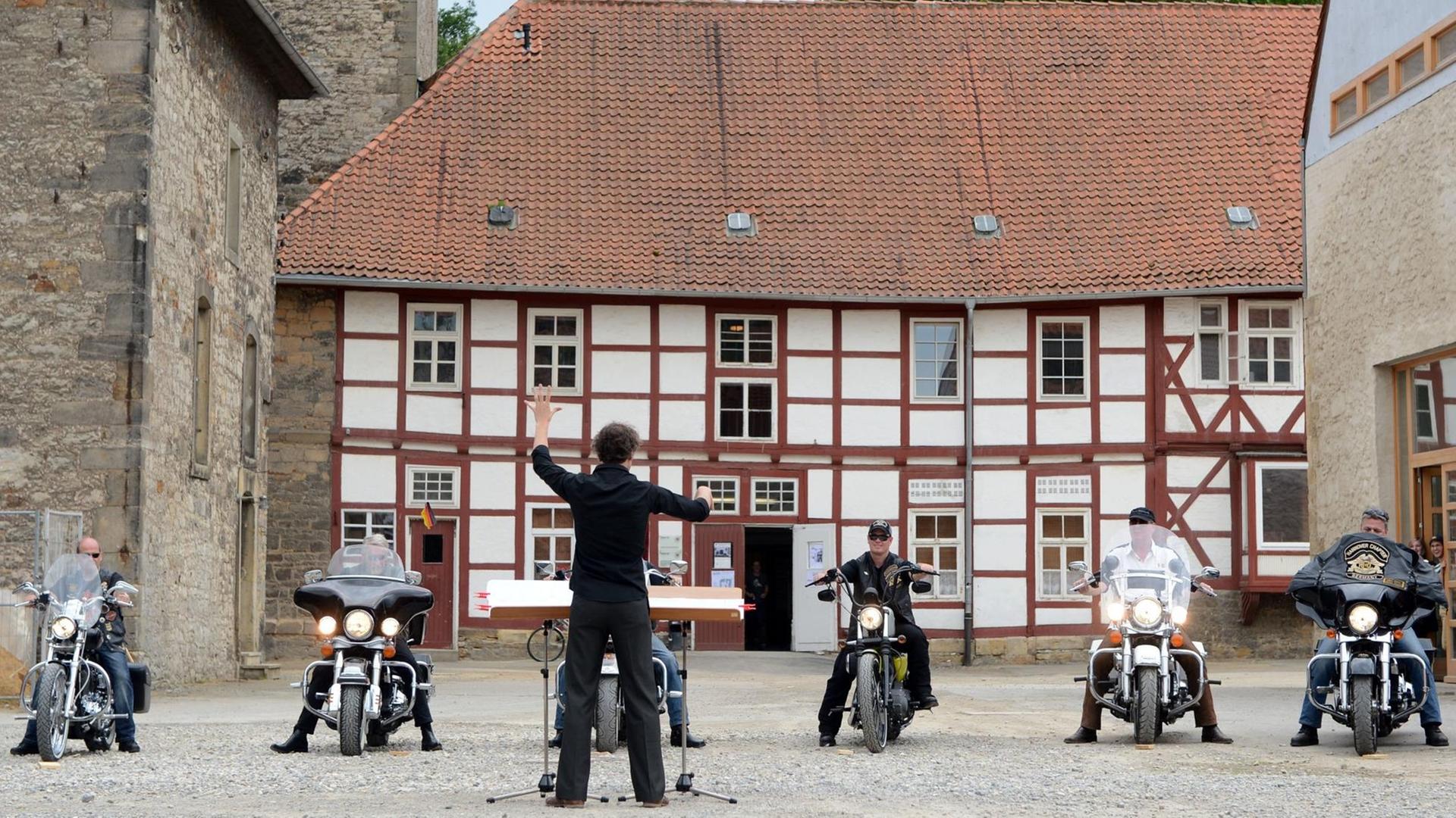 Motorradfahrer stehen mit ihren Harley Davidson Bikes vor dem Dirigenten Matthias Rebstock auf dem Kulturcampus Domäne Marienburg in Hildesheim. Der Dirigent steht mit dem Rücken zur Kamera im Zentrum des Bildes, die Motorradfahrer blicken in Richtung der Kamera, im Hintergrund ist ein Fachwerkhaus zu sehen.