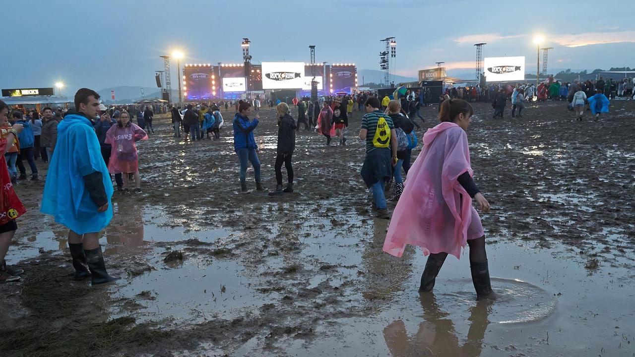 Festivalbesucher mit Regenjacken waten durch den Schlamm, im Hintergrund die beleuchtete Bühne. Festivalbesucher mit Regenjacken waten durch den Schlamm, im Hintergrund die beleuchtete Bühne.