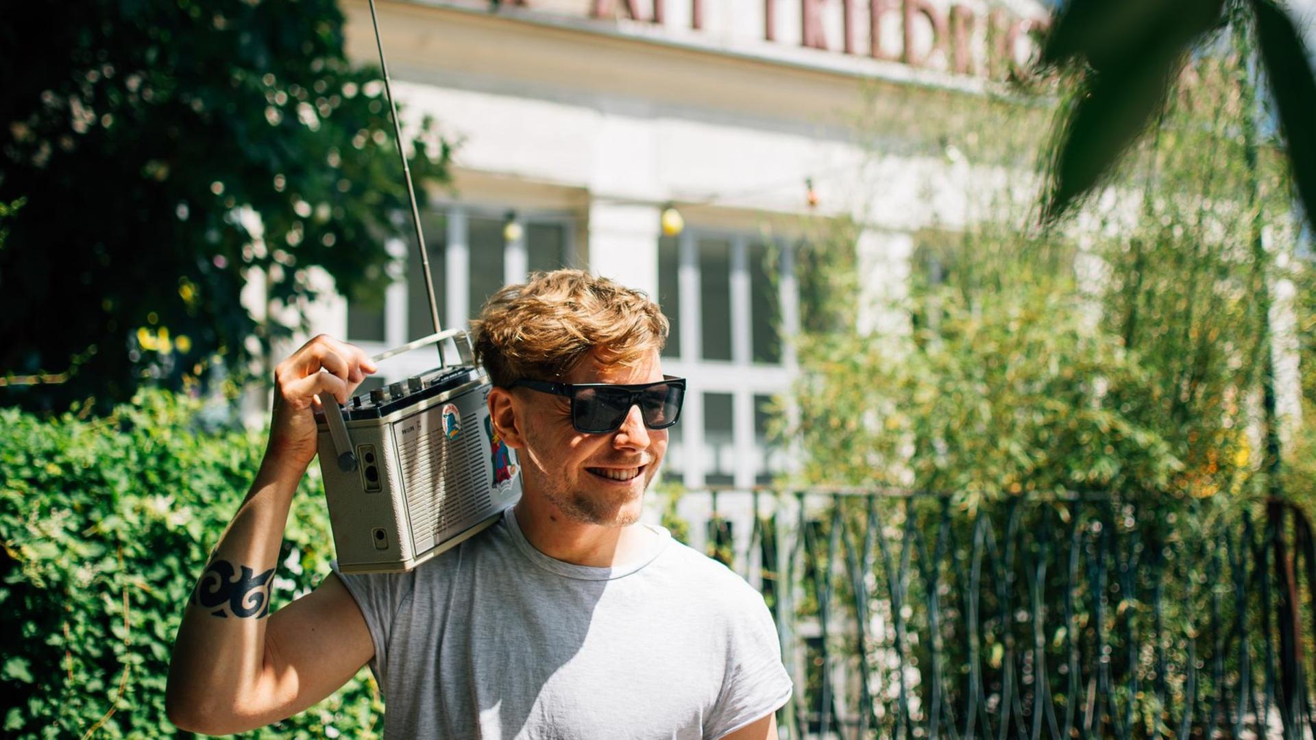 Ein junger blonder Mann mit Sonnenbrille hält ein Radiogerät mit ausgezogener Antenne auf der rechten Schulter. Ein junger blonder Mann mit Sonnenbrille hält ein Radiogerät mit ausgezogener Antenne auf der rechten Schulter.