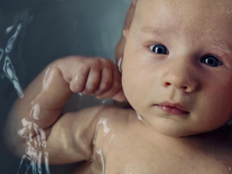 Ein Baby wird gebadet und schaut geradewegs in die Kamera. Um den Kopf ist eine schützende Hand gelegt. (Symbolfoto)