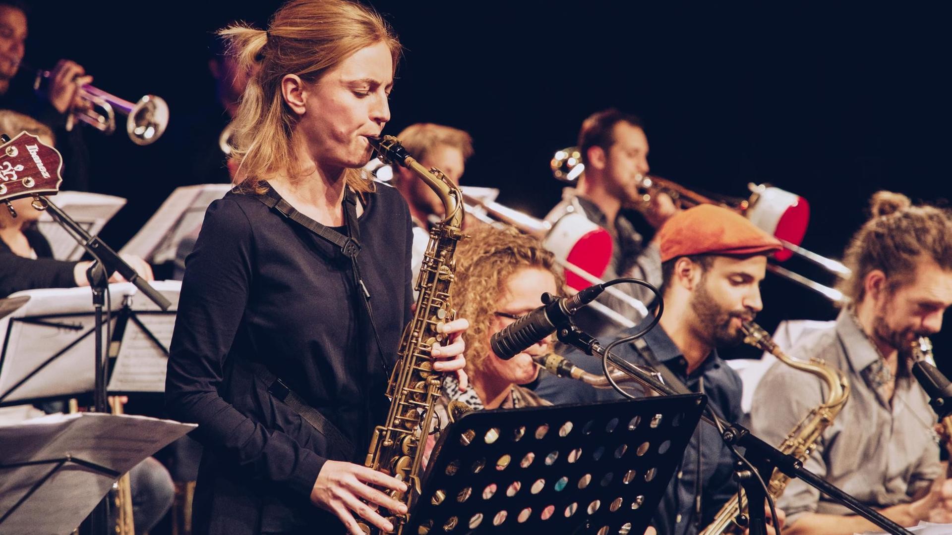 Im Vordergrund ist eine Altsaxofon spielende Frau zu sehen. Sie trägt ein schwarzes Oberteil und steht vor einem Notenständer. Im Hintergrund spielt ein Orchester auf verschiedene Blasinstrumenten Im Vordergrund ist eine Altsaxofon spielende Frau zu sehen. Sie trägt ein schwarzes Oberteil und steht vor einem Notenständer. Im Hintergrund spielt ein Orchester auf verschiedene Blasinstrumenten