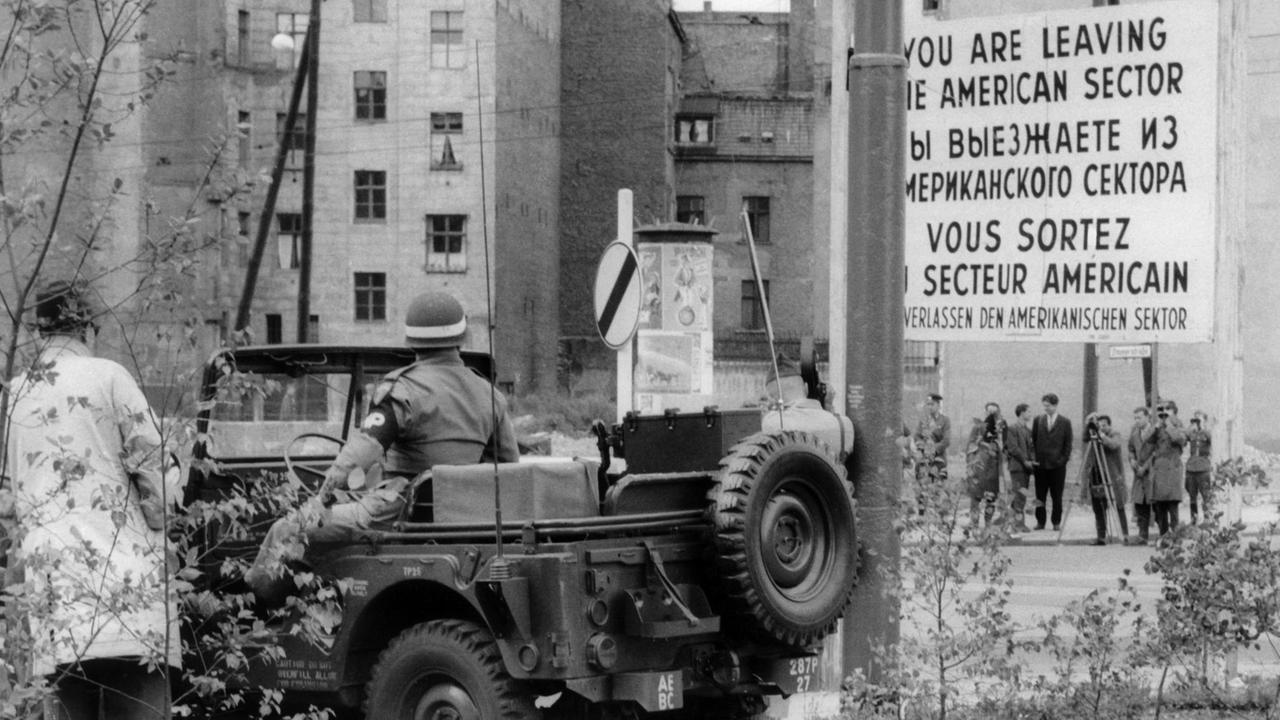 Ein Soldat der US-Armee in einem Jeep auf Westberliner und Passanten und Volkspolizisten der DDR auf Ostberliner Seite stehen sich am 23.08.1961 am Grenzübergang Friedrichstraße in Berlin gegenüber. 