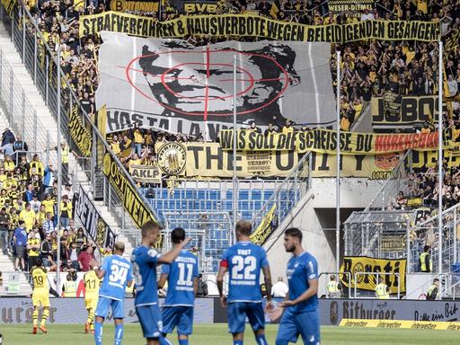 Man sieht das Spielfeld im Stadion der TSG Hoffenheim. Im Hintergrund haben BVB-Fans ein großes Banner mit dem Konterfei von Hoffenheim-Mäzen Dietmar Hopp mit einem Fadenkreuz entrollt.