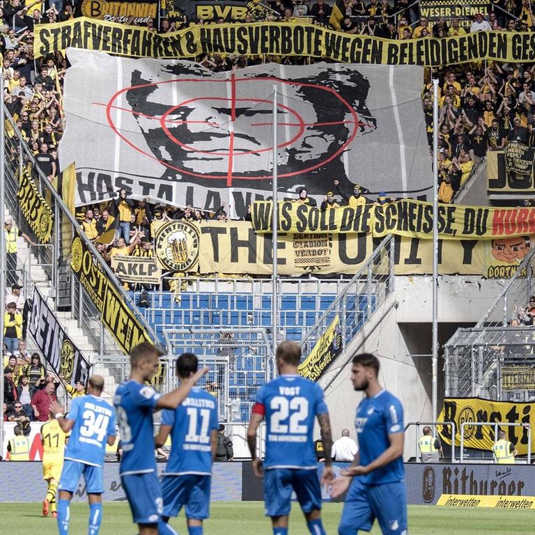 Man sieht das Spielfeld im Stadion der TSG Hoffenheim. Im Hintergrund haben BVB-Fans ein großes Banner mit dem Konterfei von Hoffenheim-Mäzen Dietmar Hopp mit einem Fadenkreuz entrollt.