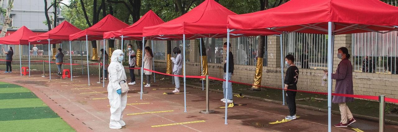 Vor einer Grundschule stehen Menschen mit Abstand in einer Warteschlange unter roten Pavillon-Dächern. Daneben ein Helfer in weißer Schutzkleidung. Vor einer Grundschule stehen Menschen mit Abstand in einer Warteschlange unter roten Pavillon-Dächern. Daneben ein Helfer in weißer Schutzkleidung.