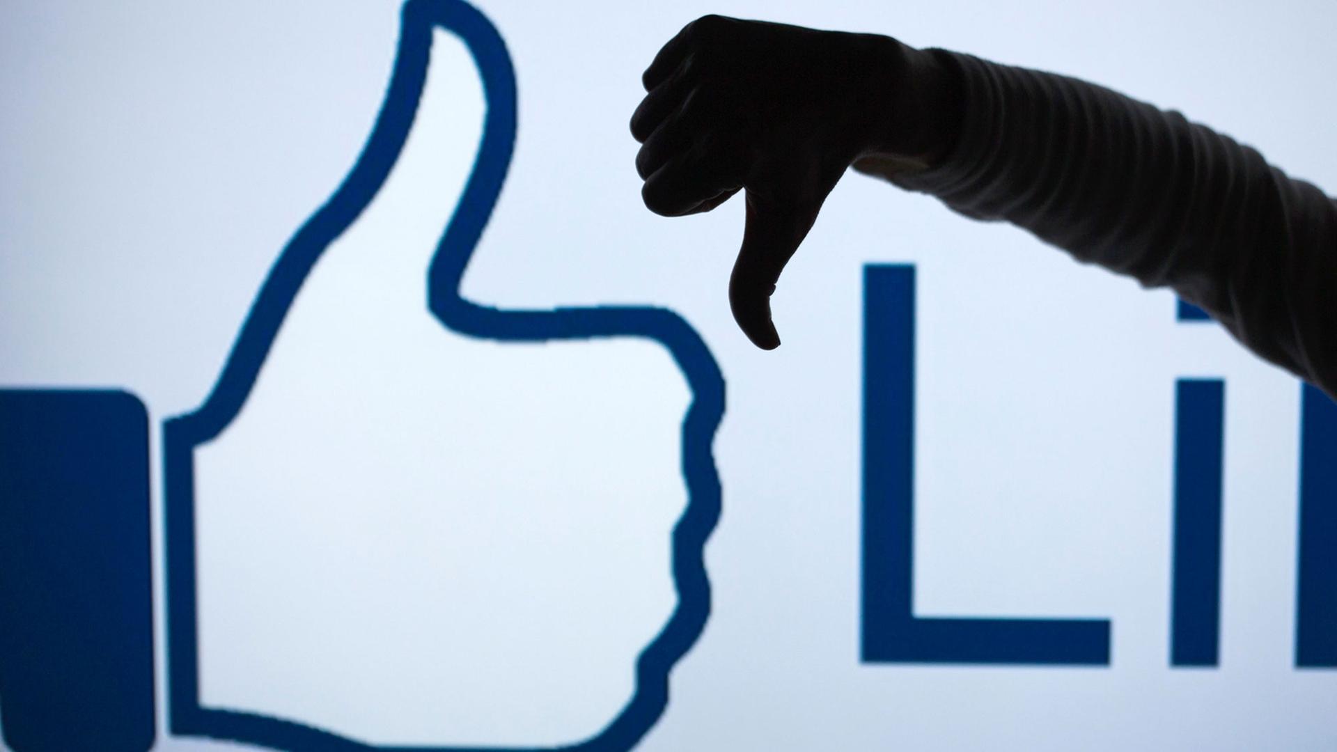 Ein Mann zeigt vor dem vergrößerten Like-Symbol der Facebook-Seite mit seinem Daumen nach unten (gestelltes Foto). Ein Mann zeigt vor dem vergrößerten Like-Symbol der Facebook-Seite mit seinem Daumen nach unten (gestelltes Foto).