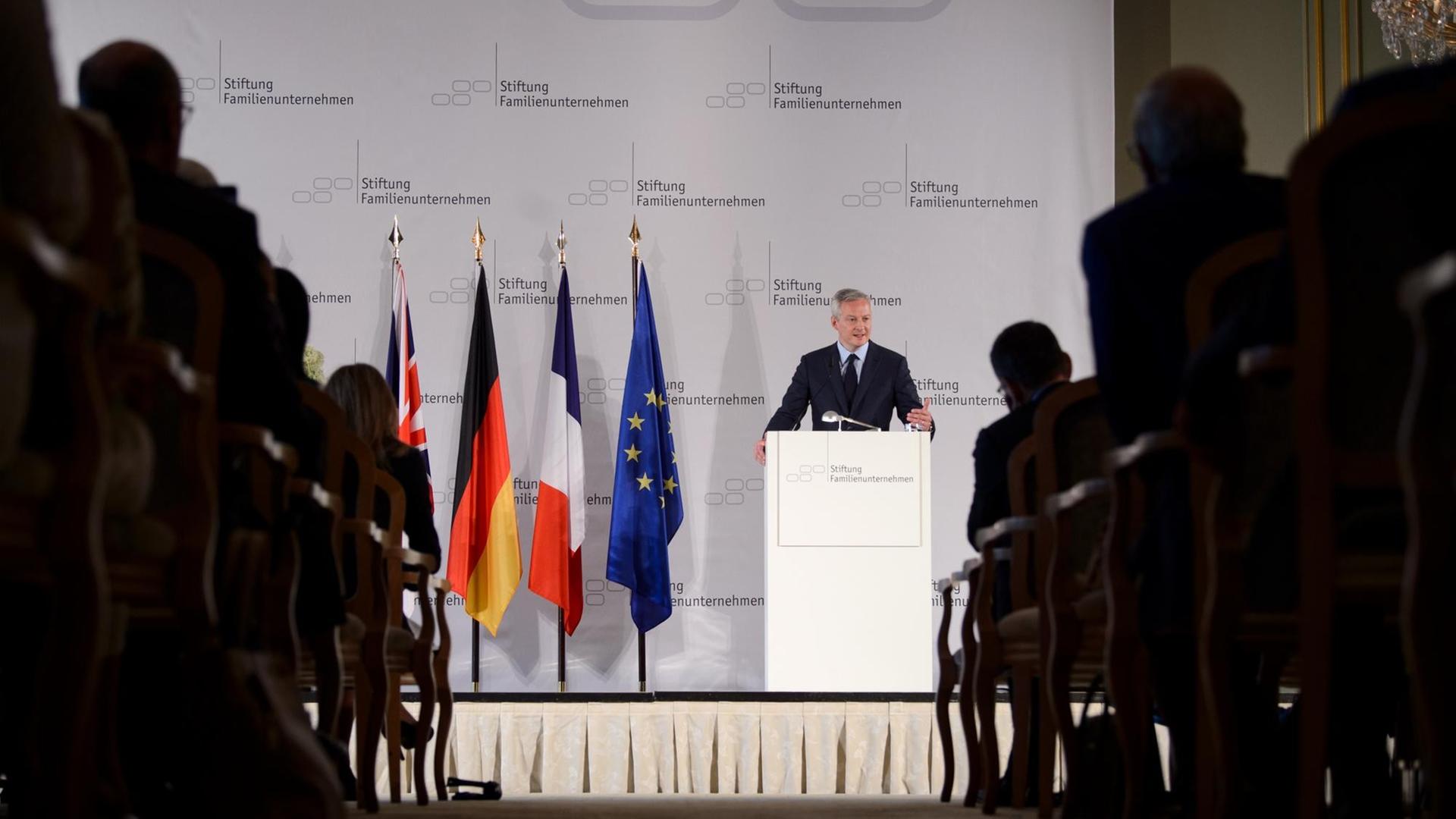 Das Bild zeigt aus größerer Entfernung Bruno Le Maire, Finanzminister von Frankreich. Er spricht beim Tag des deutschen Familienunternehmens der Stiftung für Familienunternehmen. Er steht an einem Rednerpult und hält sich daran mit beiden Händen fest.