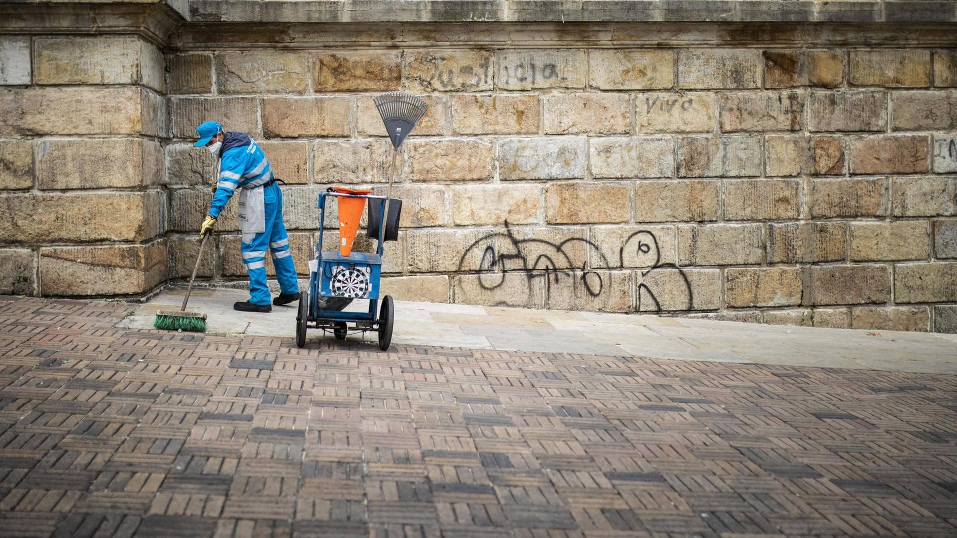 Eine Reinigungskraft in einem Blaumann schrubbt Pflaster vor einem Steingebäude Eine Reinigungskraft in einem Blaumann schrubbt Pflaster vor einem Steingebäude