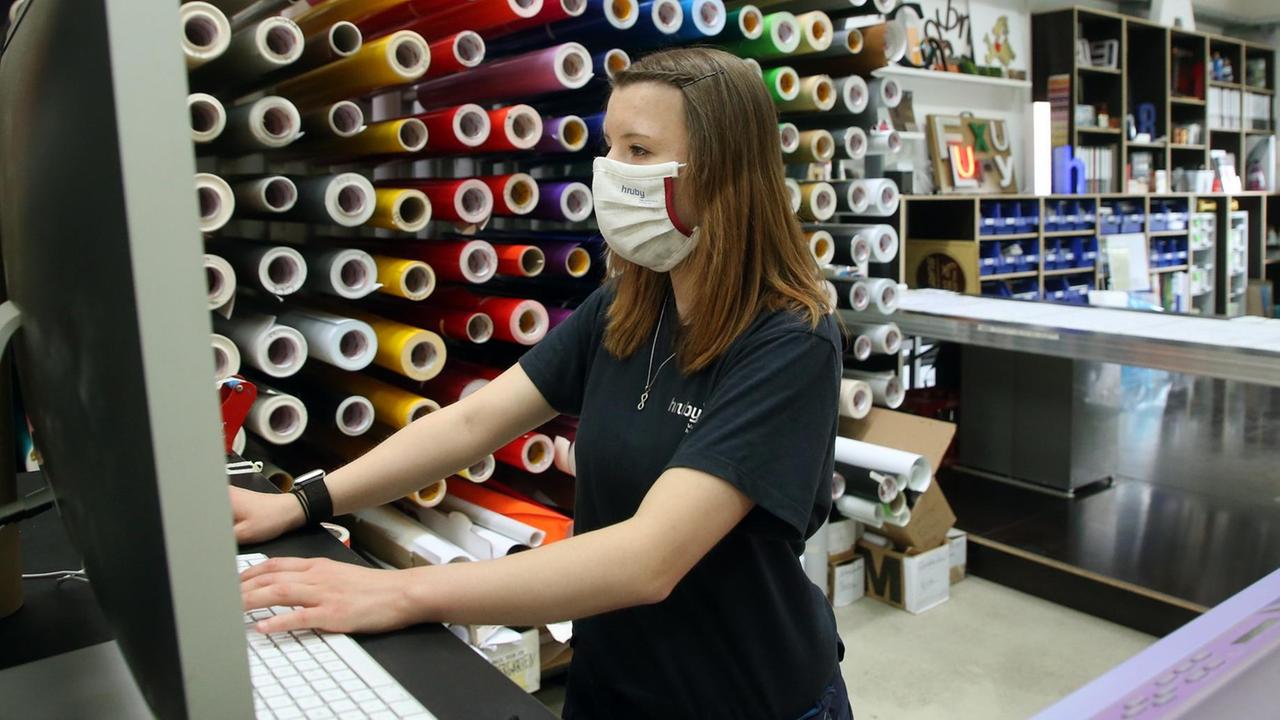 Junge Erwachsene mit Atemschutzmaske arbeitet stehend an einem Tisch in einer Werbefirma