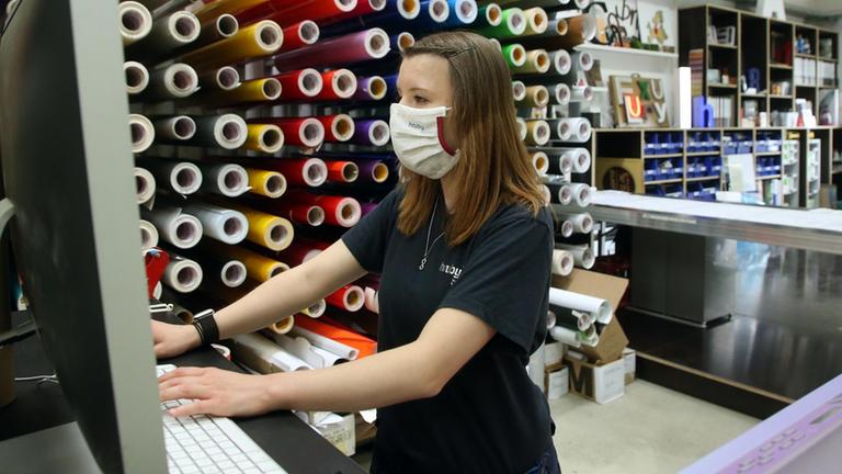 Junge Erwachsene mit Atemschutzmaske arbeitet stehend an einem Tisch in einer Werbefirma