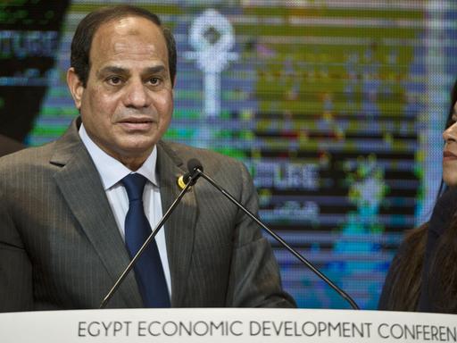 Der ägyptische Präsident Abdelfatah al-Sisi spricht an einem Rednerpult. Hinter ihm ist ein bunte TV-Leinwand zu sehen. Am rechten Rand blickt ihn eine Frau mit langen schwarzen Haaren an.