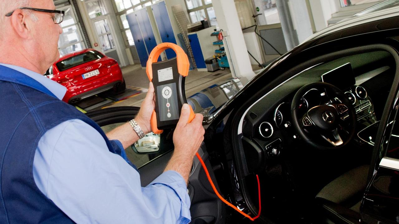 Das Bild zeigt einen älteren blaugekleideten TÜV-Mitarbeiter mit einem HU-Adapter an der öffenen Tür eines Autos. Das Bild zeigt einen älteren blaugekleideten TÜV-Mitarbeiter mit einem HU-Adapter an der öffenen Tür eines Autos.