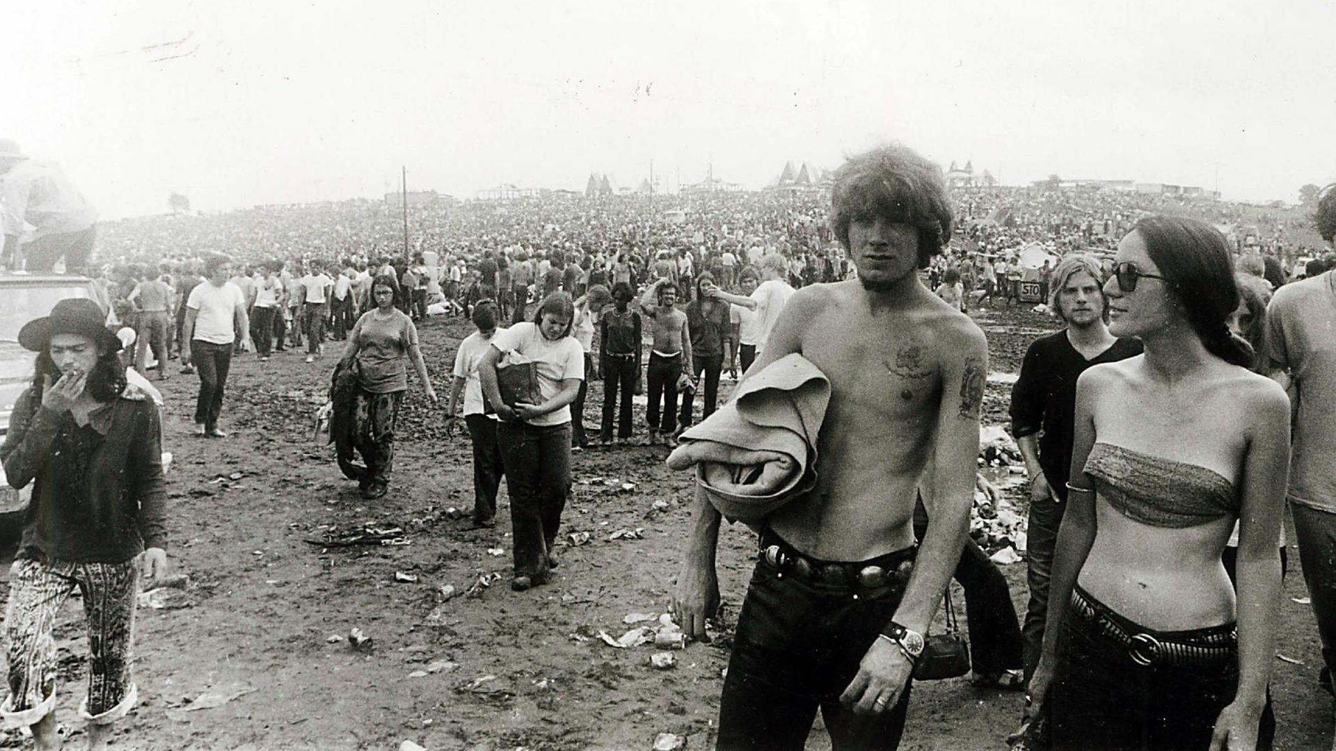 Woodstock 1969, schwarz-weiß Fotografie: Festivalbesucher laufen über den schlammigen Boden. Rechts eine junge Frau mit Sonnenbrille und ein Mann ohne T-Shirt.