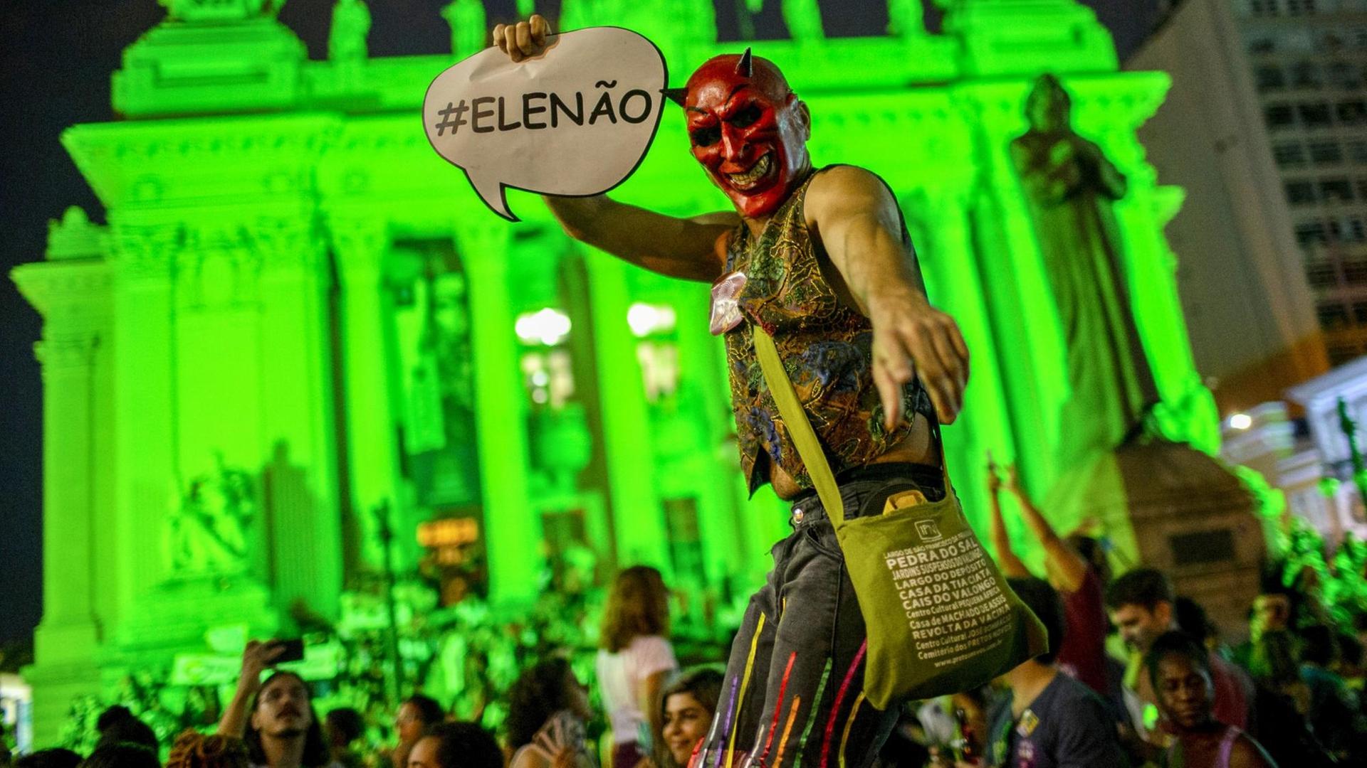 Ein Demonstrant mit Teufelsmaske hält ein Plakat mit "#EleNao" in die Kamera vor einem neongrün illuminierten historischen Gebäude.