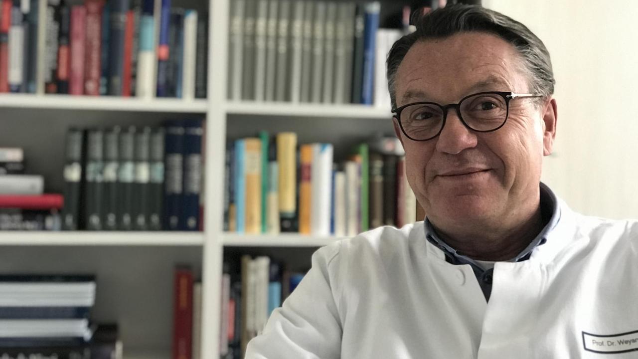 Der Herzchirurg Michael Weyand sitzt im Arztkittel vor einem Bücherregal. Der Herzchirurg Michael Weyand sitzt im Arztkittel vor einem Bücherregal.
