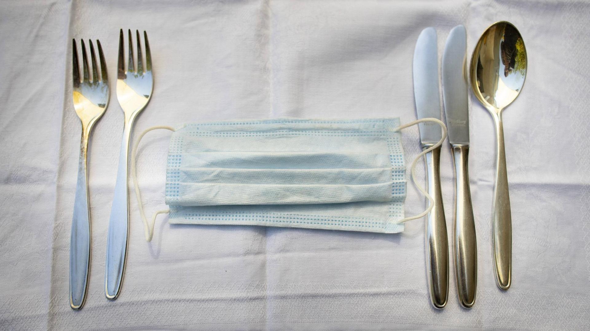 Ein medizinischer Mundschutz zwischen Messer und Gabel auf einer Tischdecke im Restaurant. Ein medizinischer Mundschutz zwischen Messer und Gabel auf einer Tischdecke im Restaurant.