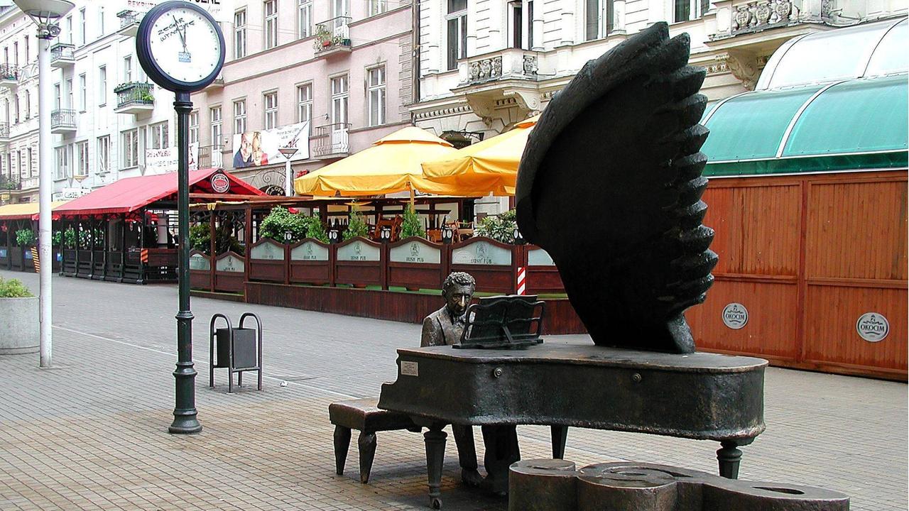 Denkmal für den Pianisten Arthur Rubinstein, der an einem großen Flügel sitzt, wobei aus dem Instrument ein Engelsflügel erwächst. Davor liegt ein massiver Notenschlüssel auf dem gepflasterten Boden.