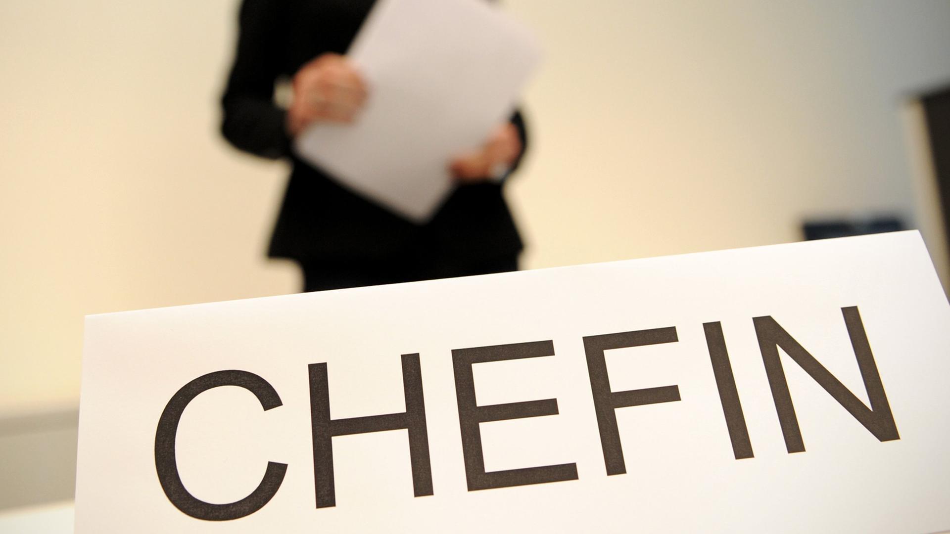 Ein Schild mit der Aufschrift "Chefin". Verschwommen im Hintergrund: Eine Frau in Business-Kleidung, die Papier in Händen hält. Ein Schild mit der Aufschrift "Chefin". Verschwommen im Hintergrund: Eine Frau in Business-Kleidung, die Papier in Händen hält.