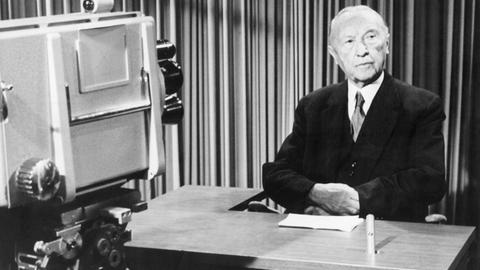Bundeskanzler Konrad Adenauer sitzt während der Aufzeichnung einer Fernsehansprache in einem Fernsehstudio hinter einem Tisch.