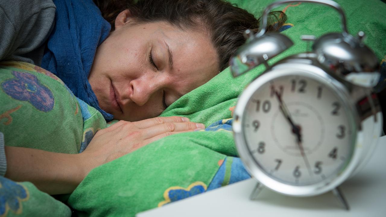 Eine Frau schläft im Bett, während ein Wecker neben ihr auf dem Nachttischränkchen steht. Eine Frau schläft im Bett, während ein Wecker neben ihr auf dem Nachttischränkchen steht.