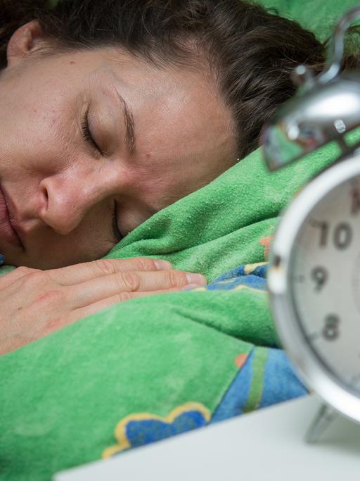 Eine Frau schläft im Bett, während ein Wecker neben ihr auf dem Nachttischränkchen steht.