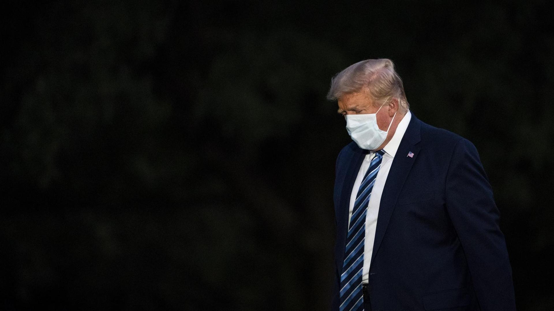 Donald Trump trägt eine Maske und steht mit geneigtem Kopf vor einem schwarzen Hintergrund Donald Trump trägt eine Maske und steht mit geneigtem Kopf vor einem schwarzen Hintergrund