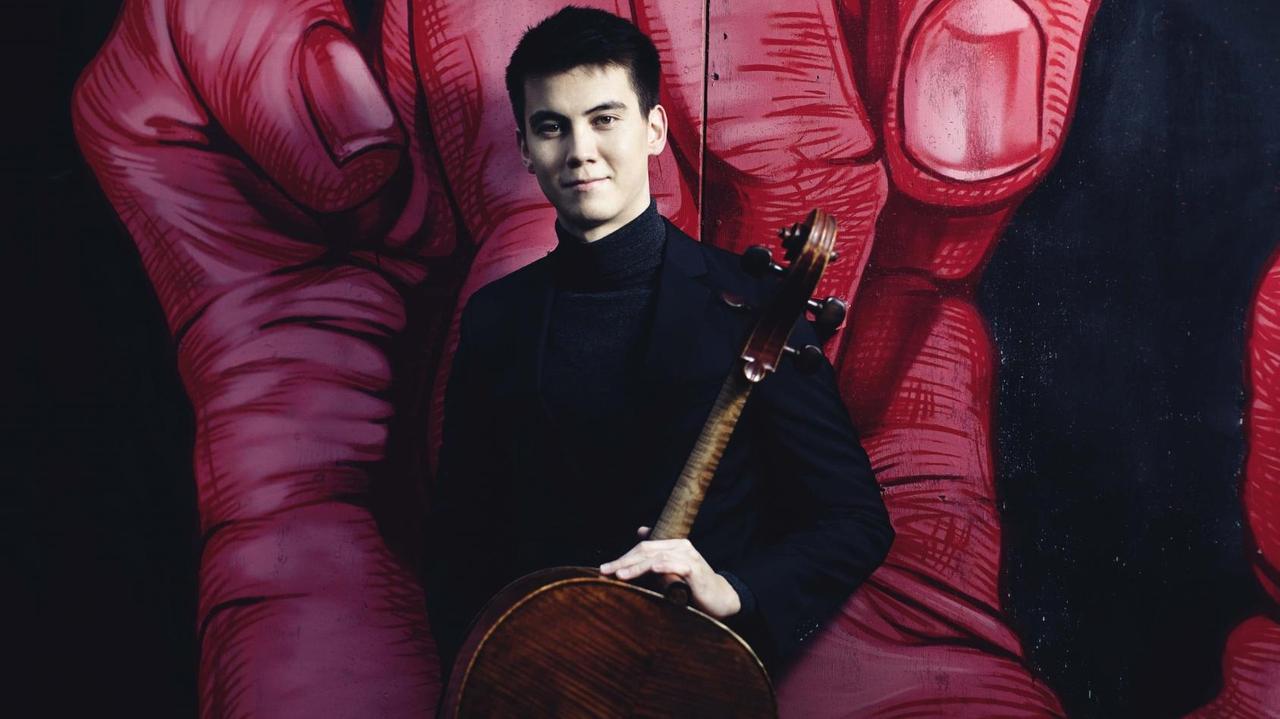 Der junge Cellist mit schwarzen, kurzen Haaren steht mit seinem Instrument vor einem Graffiti, das eine große, rote Hand mit gekrümmten Fingern zeigt. Der junge Cellist mit schwarzen, kurzen Haaren steht mit seinem Instrument vor einem Graffiti, das eine große, rote Hand mit gekrümmten Fingern zeigt.