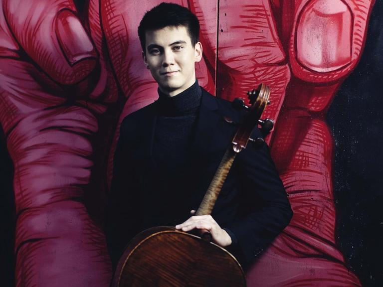 Der junge Cellist mit schwarzen, kurzen Haaren steht mit seinem Instrument vor einem Graffiti, das eine große, rote Hand mit gekrümmten Fingern zeigt.