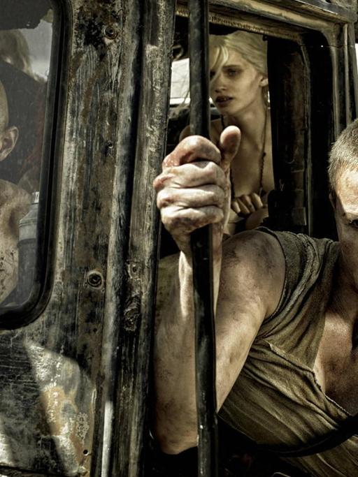 Szenenfoto aus dem Film Mad Max von 2015. Rechts Charlize Theron, die aus einem ramponierten, grauen Gefährt herausschaut.
