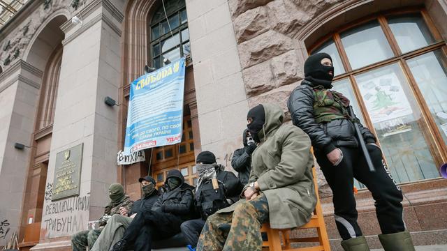 Vor dem Rathaus von Kiew sitzen Aktivisten mit schwarzen Skihauben auf Stühlen.