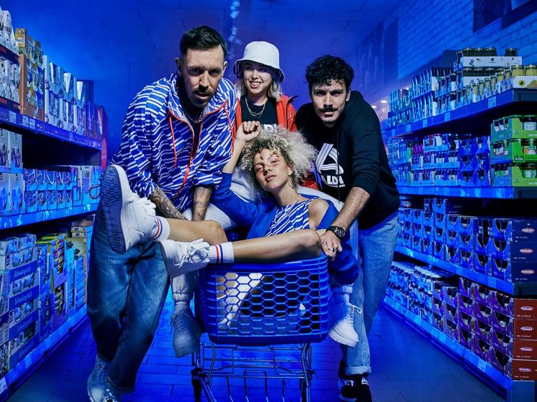 Zwei junge Männer und eine Frau stehen im Gang eines ALDI Supermarktes, in ihrer Mitte sitzt eine weitere Frau im Einkaufswagen.