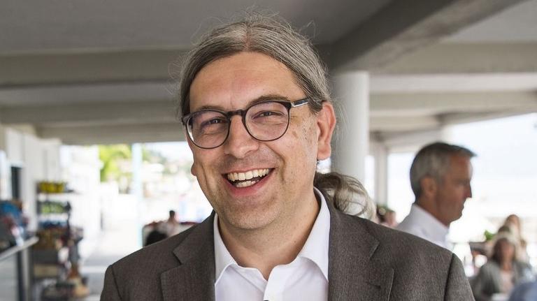 Pfarrer Thomas Zeitler, mit Brille, im offenen weißen Hemd und grauem Sakko, das lange graue Haar im Nacken zusammengebunden, lächelt in die Kamera, in seinem Rücken sind weitere Personen in einer Kantine zu sehen, aus dem Garten fällt Licht durch die großen Fenster. Pfarrer Thomas Zeitler, mit Brille, im offenen weißen Hemd und grauem Sakko, das lange graue Haar im Nacken zusammengebunden, lächelt in die Kamera, in seinem Rücken sind weitere Personen in einer Kantine zu sehen, aus dem Garten fällt Licht durch die großen Fenster.