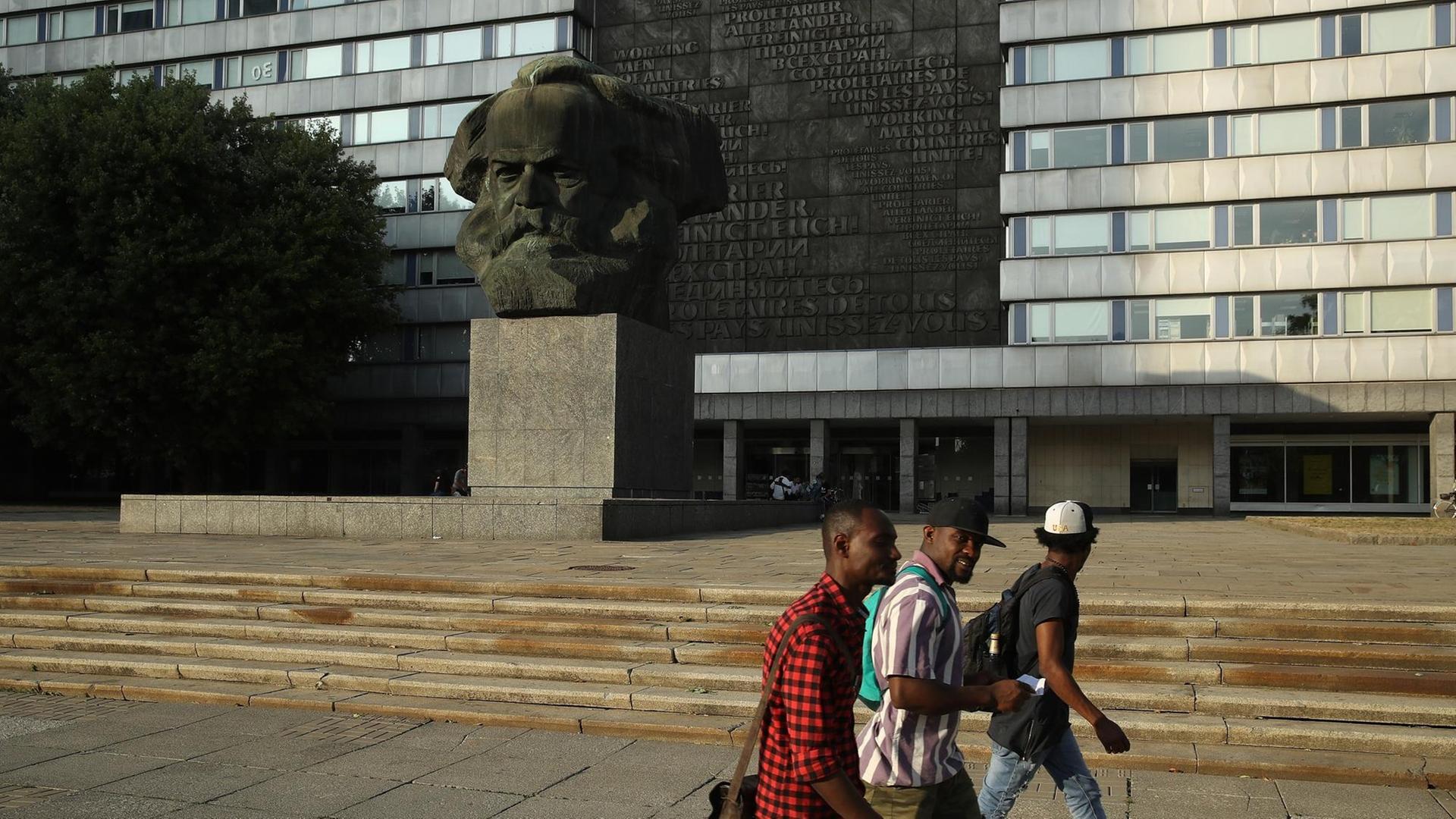 Drei schwarze Menschen laufen in Chemnitz an dem großen Karl-Marx-Kopf in der Innenstadt vorbei.