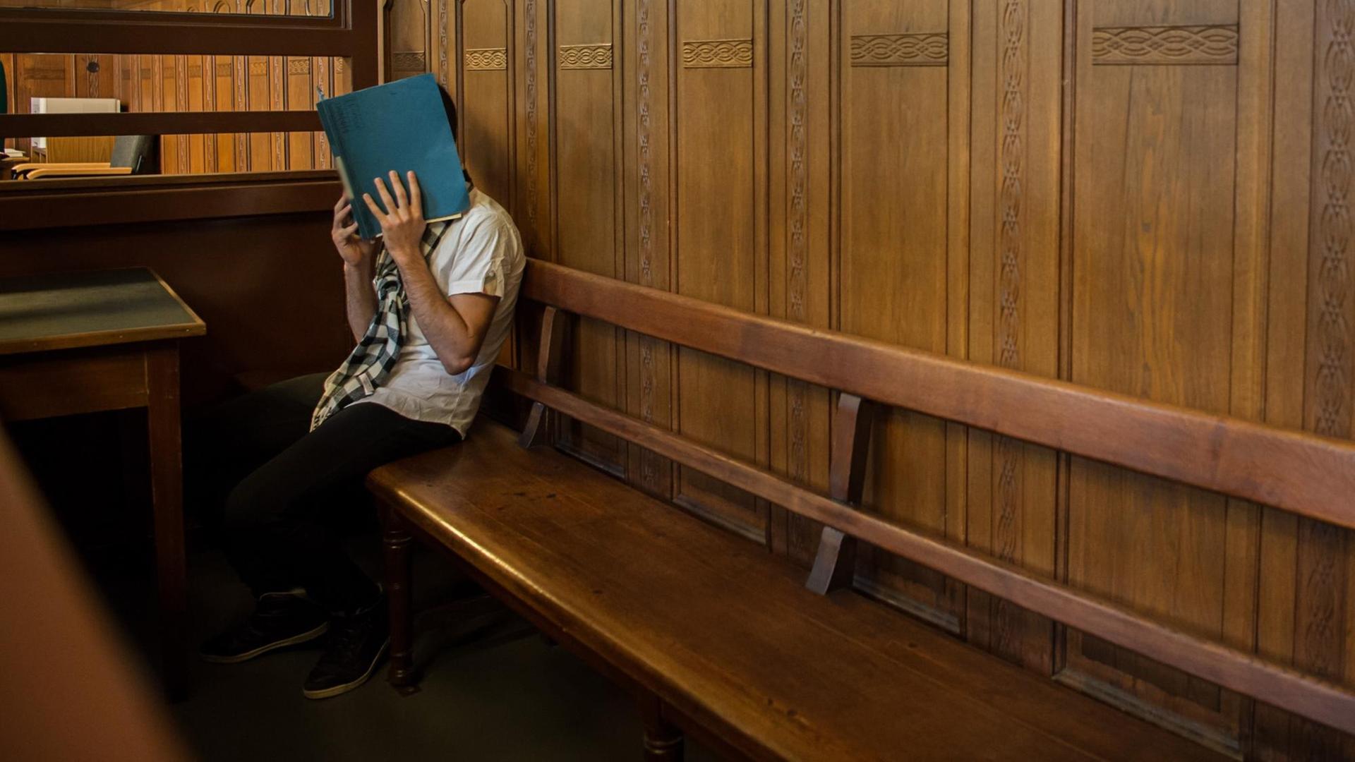 Ein Angeklageter sitzt auf einer langen hölzernen Anklagebank am Langericht Berlin-Moabit. Er verdeckt sein Gesicht mit einem Aktendeckel.
