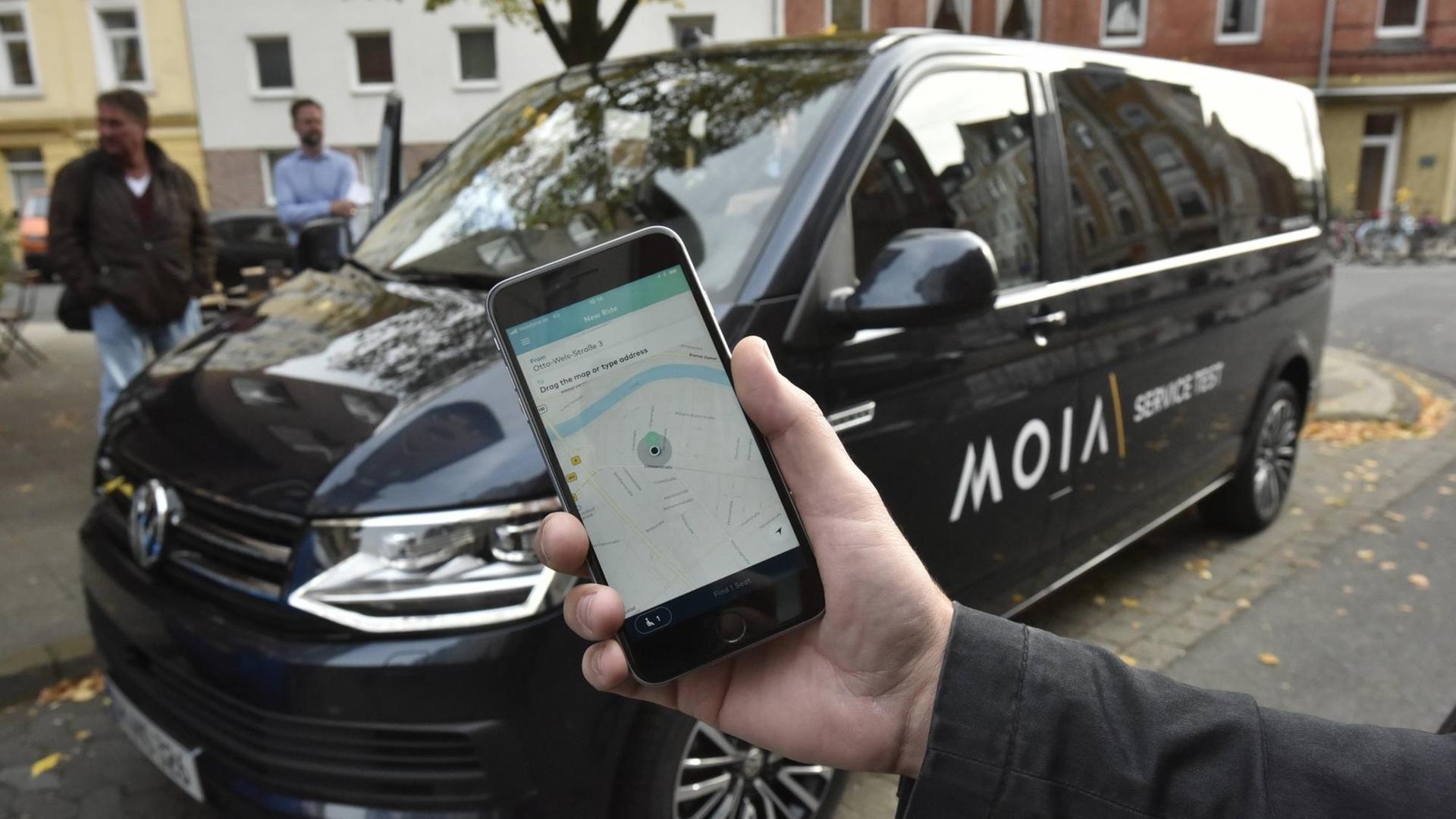 Ein Handy mit der MOIA-App vor einem Fahrzeug des Unternehmens. Ein Handy mit der MOIA-App vor einem Fahrzeug des Unternehmens.