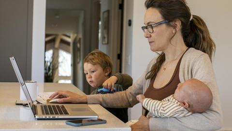 Eine Mutter im Homeoffice vor dem Laptop, mit Baby auf dem Schoß und einem spielenden Kleinkind neben sich. (Symbolbild)