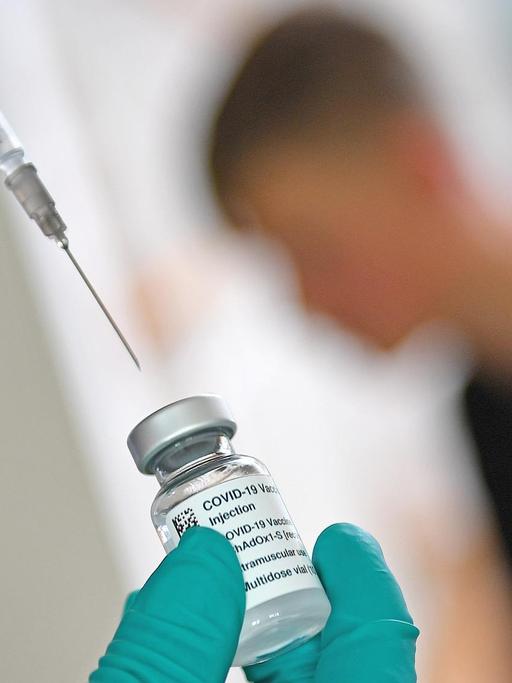 Mit einer Spritze wird Impfserum gegen Covid 19 aus einer Ampulle gezogen. Im Hintergrund ist ein Jugendlicher zu erkennen, der geimpft werden soll.
